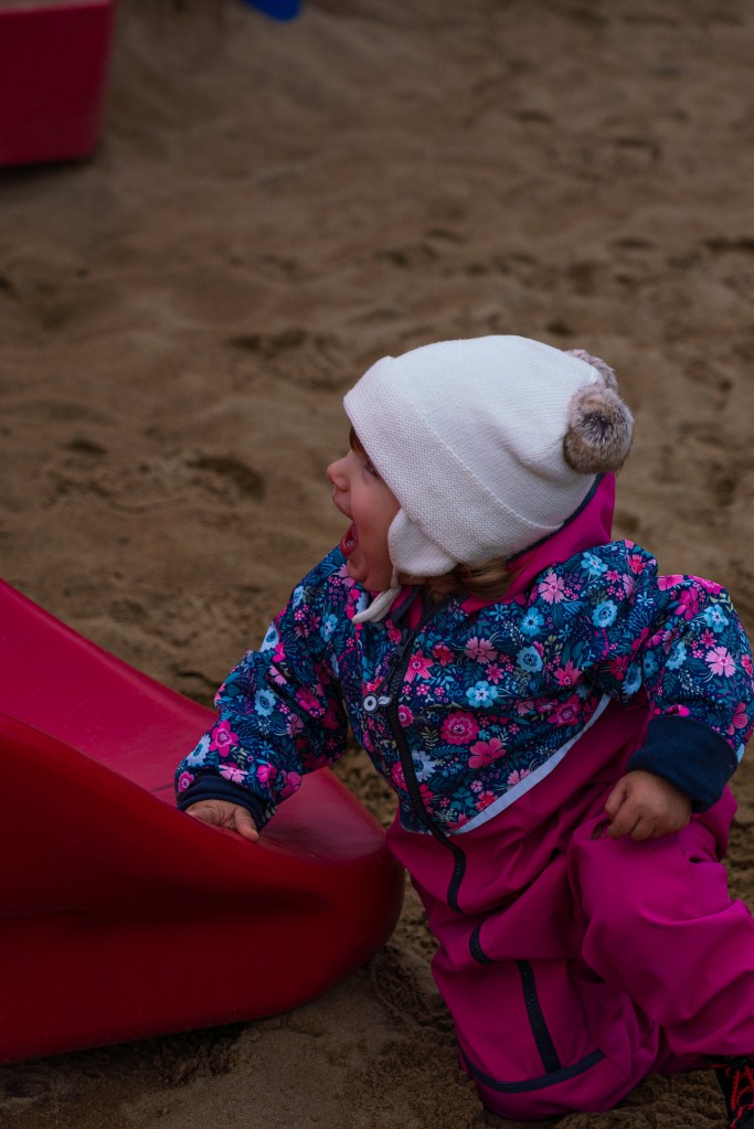 Une jeune enfant avec une combinaison colorée joue près d'un toboggan rouge sur une aire de jeux en sable, exprimant de la joie.