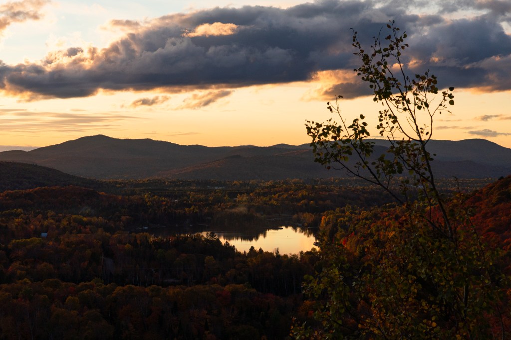 Vue panoramique sur un paysage d'automne au Canada, avec des montagnes en arrière-plan et un lac reflétant le ciel au coucher du soleil. Des arbres aux couleurs flamboyantes entourent le lac, ajoutant à la beauté de la scène.