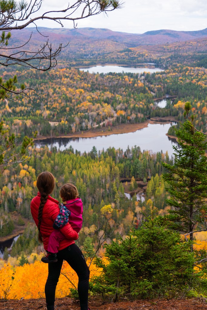 Une mère portant son enfant admire un panorama d'automne au Canada, avec des arbres aux couleurs vives et des lacs reflétant le paysage.