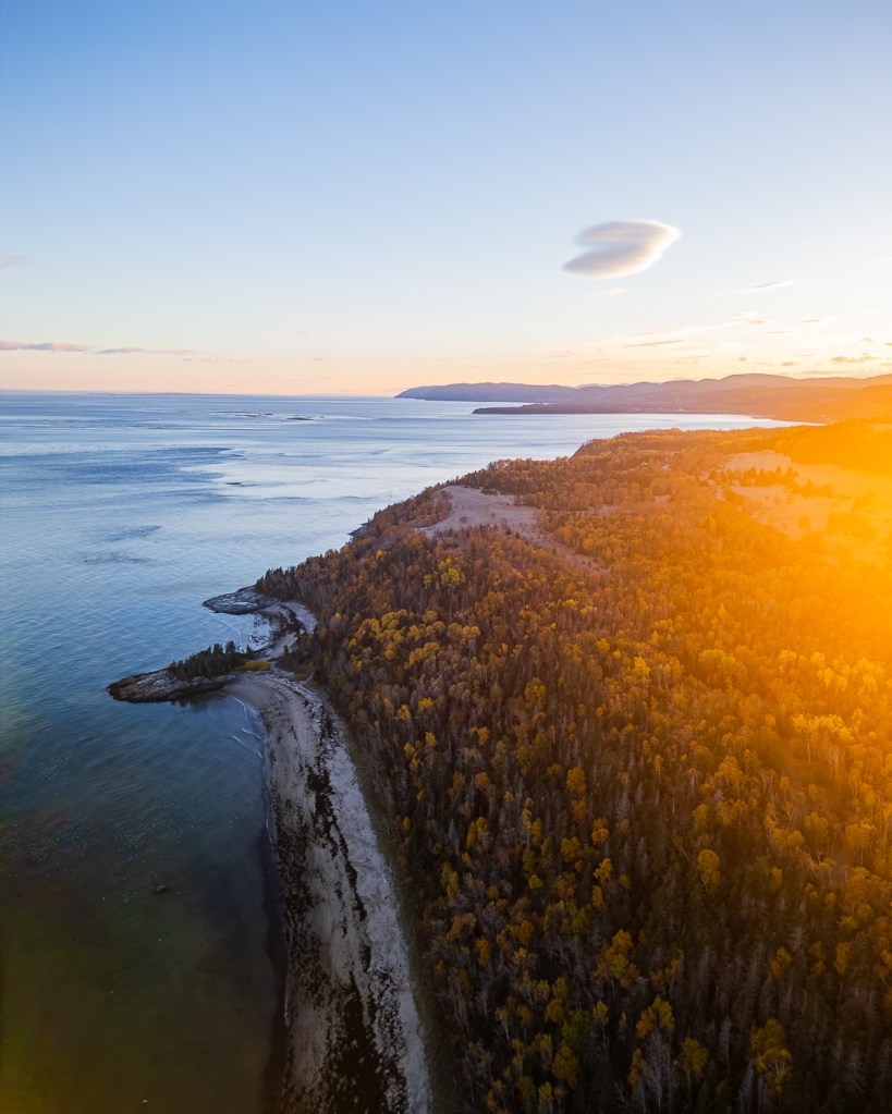 Vue aérienne panoramique sur la côte du Québec au crépuscule, avec des reflets dorés sur l'eau et une forêt aux teintes d'automne.