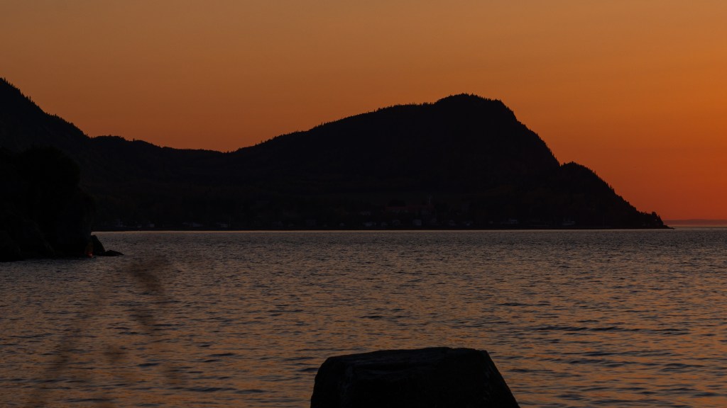 Silhouette des montagnes se profilant contre un ciel orange vif au coucher de soleil, avec un reflet sur l'eau calme.