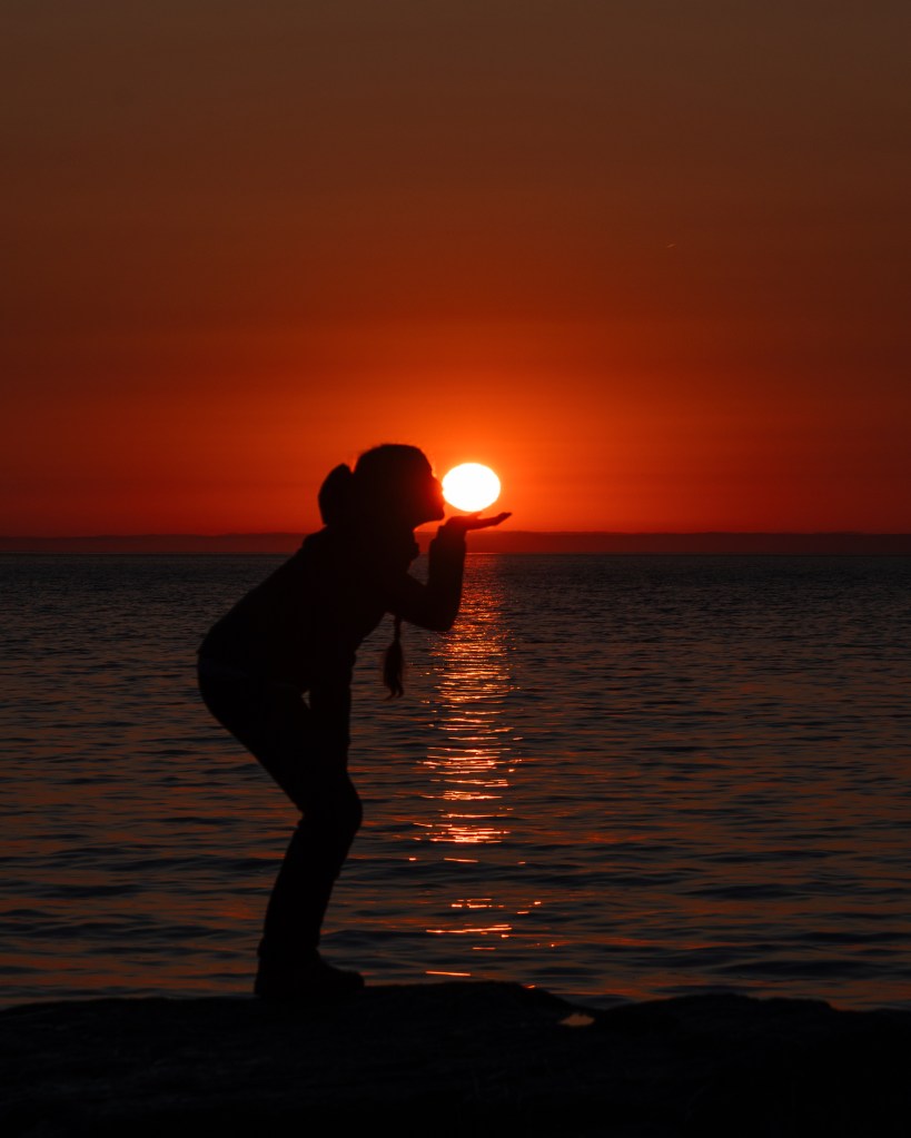 Silhouette d'une personne faisant semblant de tenir le soleil qui se lève ou se couche au-dessus de l'eau, avec des reflets dorés sur la surface de l'eau.