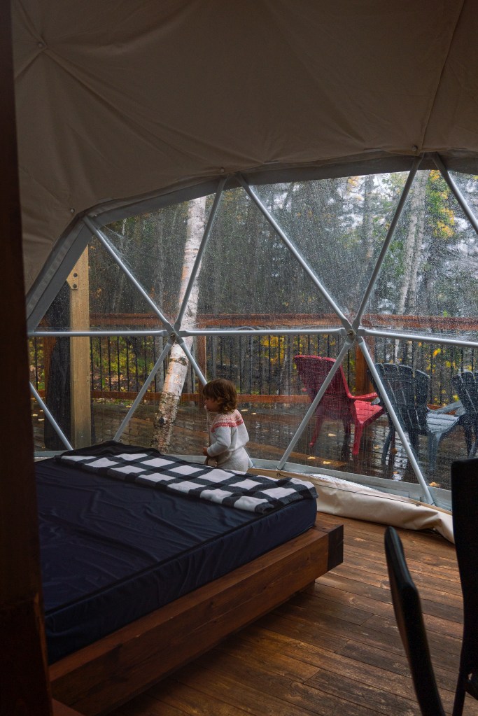 Intérieur d'un dôme en bois avec un lit et des murs en verre, montrant un enfant jouant près d'une fenêtre avec des gouttes de pluie sur le verre et un paysage forestier à l'extérieur.