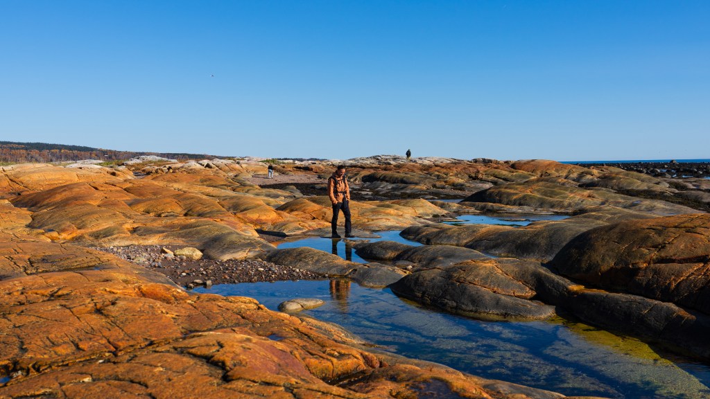 A person walking on rocky shoreline with orange and black rock formations under a clear blue sky.