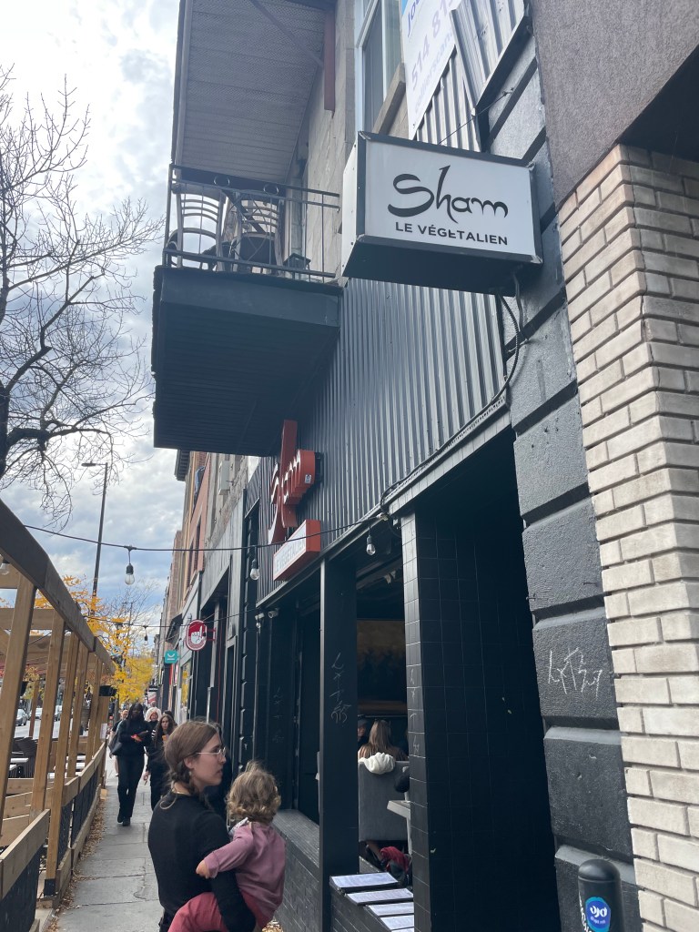 Vue extérieure du restaurant végétalien Sham avec une mère tenant un enfant sur le trottoir.