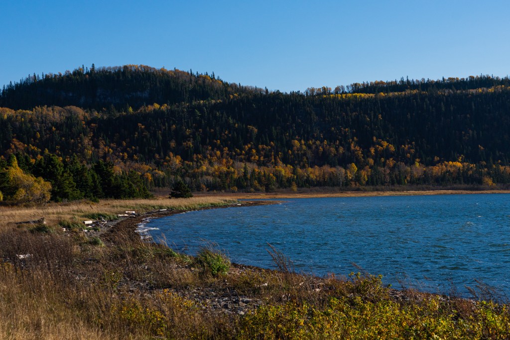 Vue d'un paysage automnal avec des collines boiséess, un ciel bleu et un lac en premier plan, soulignant les couleurs dorées et vertes des arbres.