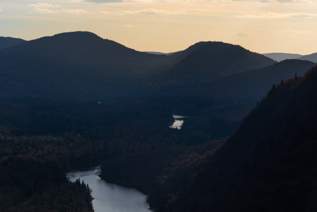 Vue panoramique sur des montagnes et une vallée avec une rivière sinueuse sous un ciel partiellement nuageux.